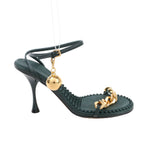 Bottega Veneta green leather heel sandal featuring a sculpted stiletto heel, ankle strap with gold buckle and hanging orb charm, gold chain front strap, and textured footbed, displayed on a white background.