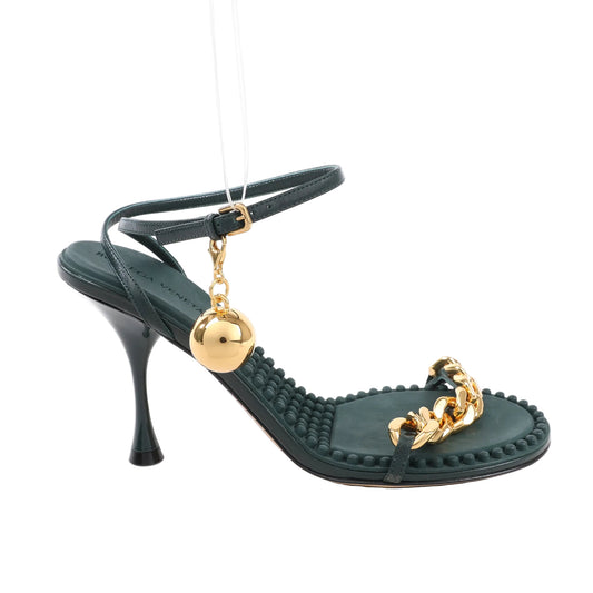 Bottega Veneta green leather heel sandal featuring a sculpted stiletto heel, ankle strap with gold buckle and hanging orb charm, gold chain front strap, and textured footbed, displayed on a white background.