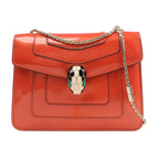BVLGARI Serpenti Forever shoulder bag in glossy orange patent leather, adorned with a gold-tone chain strap and signature snakehead clasp with black, white, and green enamel detailing, shown against a white background.