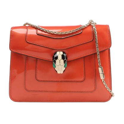 BVLGARI Serpenti Forever shoulder bag in glossy orange patent leather, adorned with a gold-tone chain strap and signature snakehead clasp with black, white, and green enamel detailing, shown against a white background.