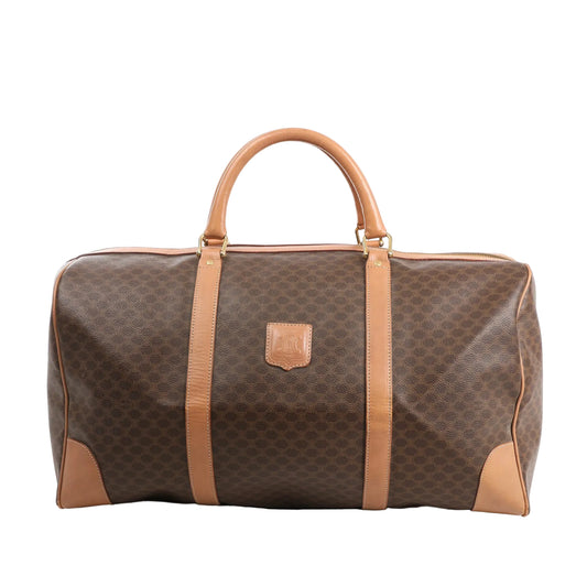 Front view of a Celine Macadam Boston travel bag in brown coated PVC with tan leather trim, dual rolled handles, gold-tone hardware, and signature logo patch.
