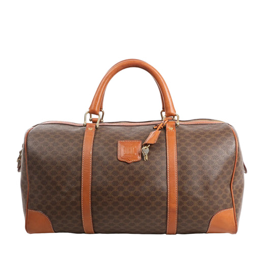 Front view of a Celine Macadam Boston travel bag in brown PVC with tan leather trim, featuring dual rolled handles, top zip closure, and signature Celine logo patch.