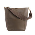 CELINE Seau Sangle Bucket Bag in greige grained leather with gold-tone hardware and wide brown fabric shoulder strap, shown standing upright against a white background.