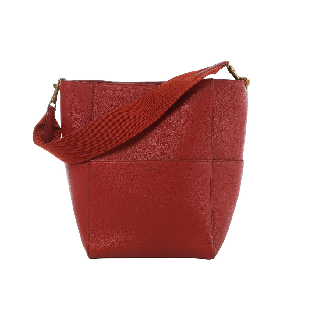 CELINE Seau Sangle bucket shoulder bag in red grained leather with a wide matching shoulder strap, gold-tone hardware and minimalist design on a white background.