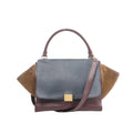 CELINE Trapeze 2Way handbag with navy blue leather flap, burgundy leather body, brown suede side wings, curved top handle, gold-tone square logo plate, and detachable shoulder strap, on a white background.