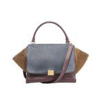 CELINE Trapeze 2Way handbag with navy blue leather flap, burgundy leather body, brown suede side wings, curved top handle, gold-tone square logo plate, and detachable shoulder strap, on a white background.