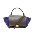 CELINE Trapeze handbag with brown leather flap, navy suede side panels, black leather base, top handle, shoulder strap, and gold-tone plaque, front view on white background.