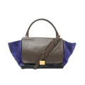 CELINE Trapeze handbag with brown leather flap, navy suede side panels, black leather base, top handle, shoulder strap, and gold-tone plaque, front view on white background.