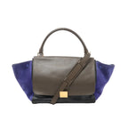 CELINE Trapeze handbag with brown leather flap, navy suede side panels, black leather base, top handle, shoulder strap, and gold-tone plaque, front view on white background.