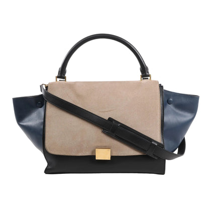 CELINE Trapeze 2Way Handbag in black, navy, and taupe, featuring a taupe suede flap, navy leather side wings, black leather body and top handle, gold-tone square clasp, and detachable black shoulder strap, displayed on a white background.