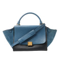 CELINE Trapeze handbag in blue leather with suede side panels and a black base, top handle and shoulder strap, front view on white background.