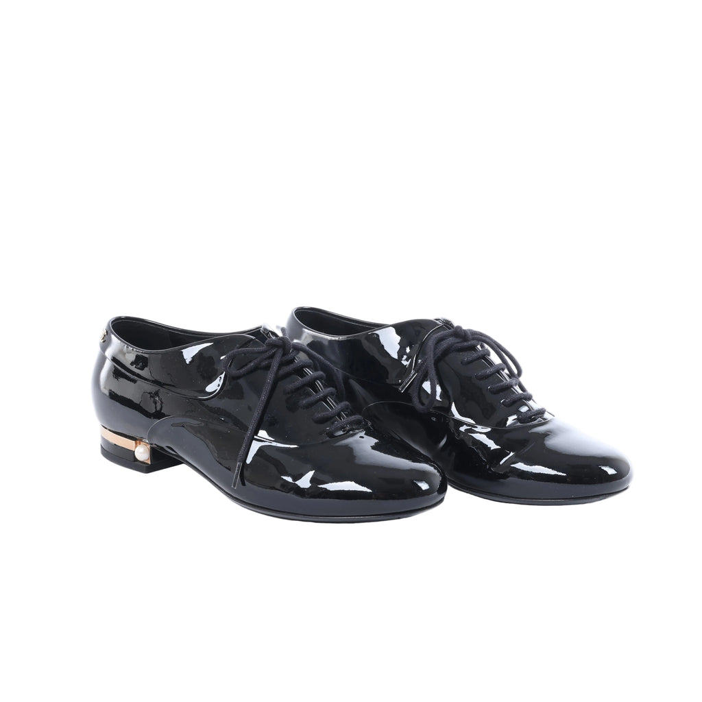 Black Chanel patent leather lace-up shoes displayed beside a black Chanel box, dust bags and white cards.