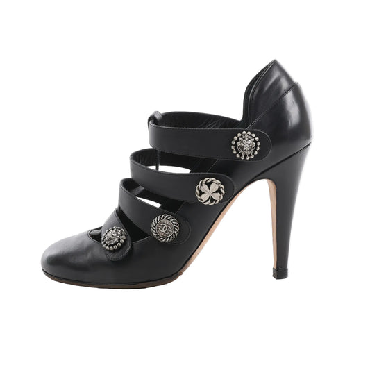 Chanel black leather pumps with high heels, featuring four front straps adorned with silver-tone buttons including the Coco mark, lion, clover, and rope designs, displayed in side profile on a white background.