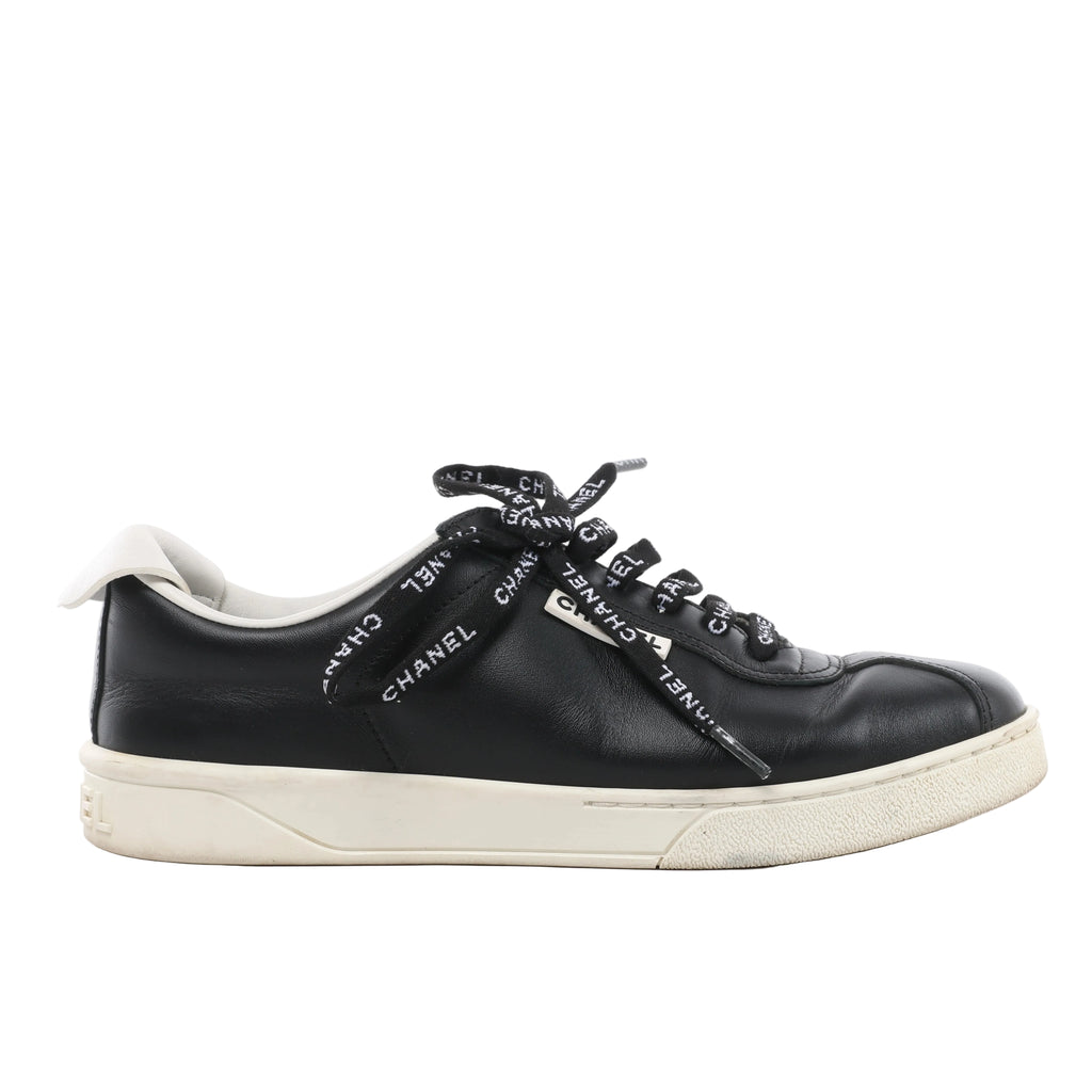 Side view of a black Chanel leather low-top sneaker with Chanel-lettered black laces, white collar trim and an off-white sole.