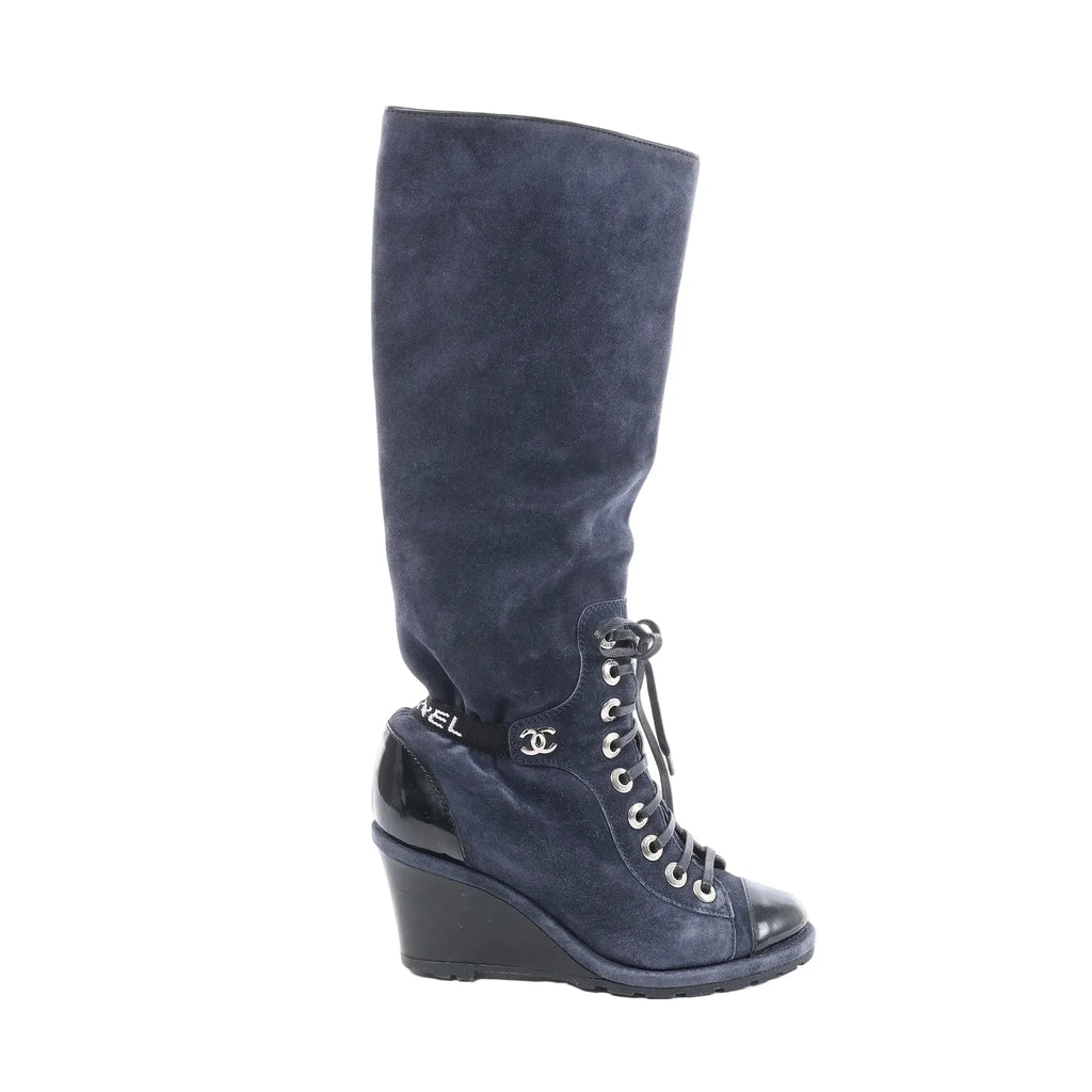 Chanel long boot in navy suede with black patent toe and wedge heel, lace-up front with silver eyelets, and black Chanel logo ankle detail, side view on white background
