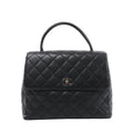 Front view of a black Chanel Kelly handbag with grained leather, diamond quilting, a curved top handle and a silver-tone CC turn-lock on the flap.