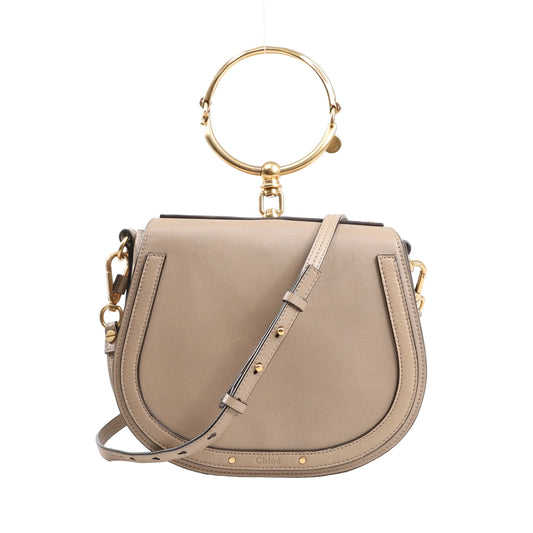Chloé Nile Small 2Way Shoulder Bag in beige leather with gold-tone metal ring handle, adjustable crossbody strap, and curved saddle silhouette on a white background.