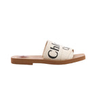 Side view of a Chloé Woody slide sandal with beige crossover fabric straps, black Chloé lettering and a flat tan sole on a white background.