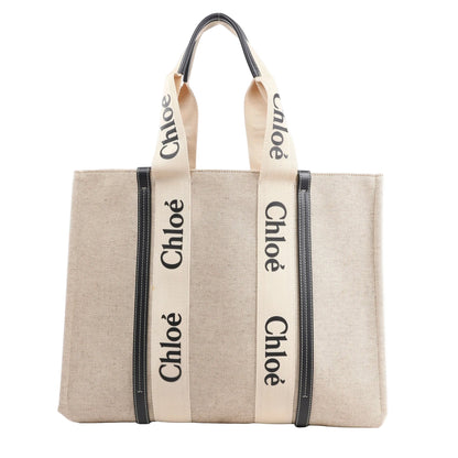 CHLOÉ Woody Large Tote bag made of beige canvas with black leather trim and wide cream fabric shoulder straps printed with the black Chloé logo, displayed upright against a white background.