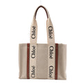 Front view of the Chloé Woody Medium Canvas & Leather Tote Shoulder Bag in brown, featuring beige canvas body, vertical brown leather trim, wide cream fabric handles with black "Chloé" logo print, displayed against a white background.