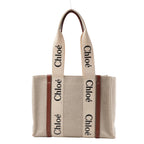 Front view of the Chloé Woody Medium Canvas & Leather Tote Shoulder Bag in brown, featuring beige canvas body, vertical brown leather trim, wide cream fabric handles with black "Chloé" logo print, displayed against a white background.