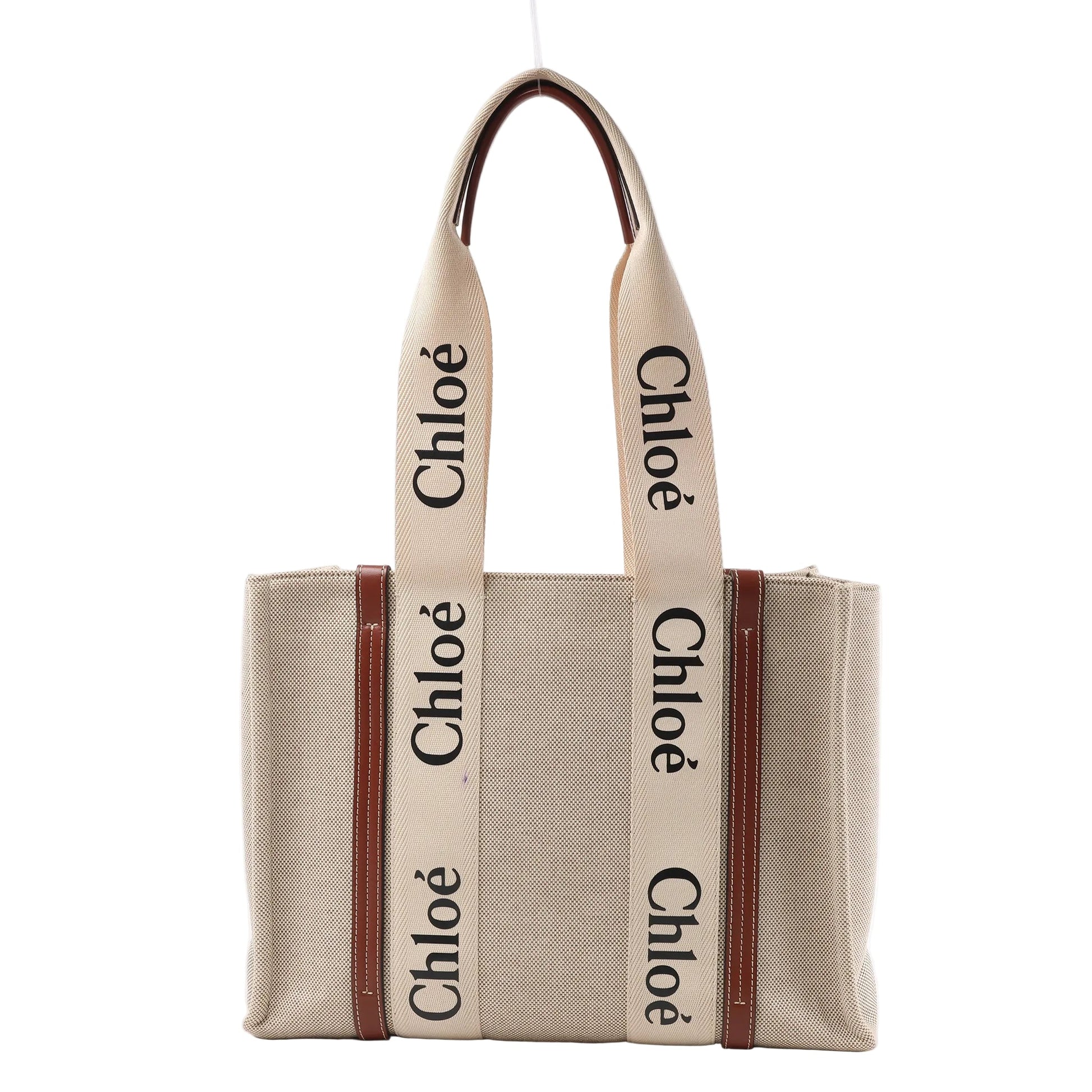 Front view of the Chloé Woody Medium Canvas & Leather Tote Shoulder Bag in brown, featuring beige canvas body, vertical brown leather trim, wide cream fabric handles with black "Chloé" logo print, displayed against a white background.