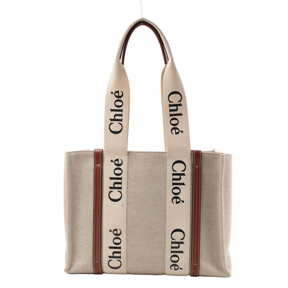 Front view of the Chloé Woody Medium Canvas & Leather Tote Shoulder Bag in brown, featuring beige canvas body, vertical brown leather trim, wide cream fabric handles with black "Chloé" logo print, displayed against a white background.