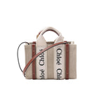Front view of Chloé Woody Nano tote in beige canvas with vertical “Chloé” logo webbing, brown leather trim, top handles, and a brown leather shoulder strap.