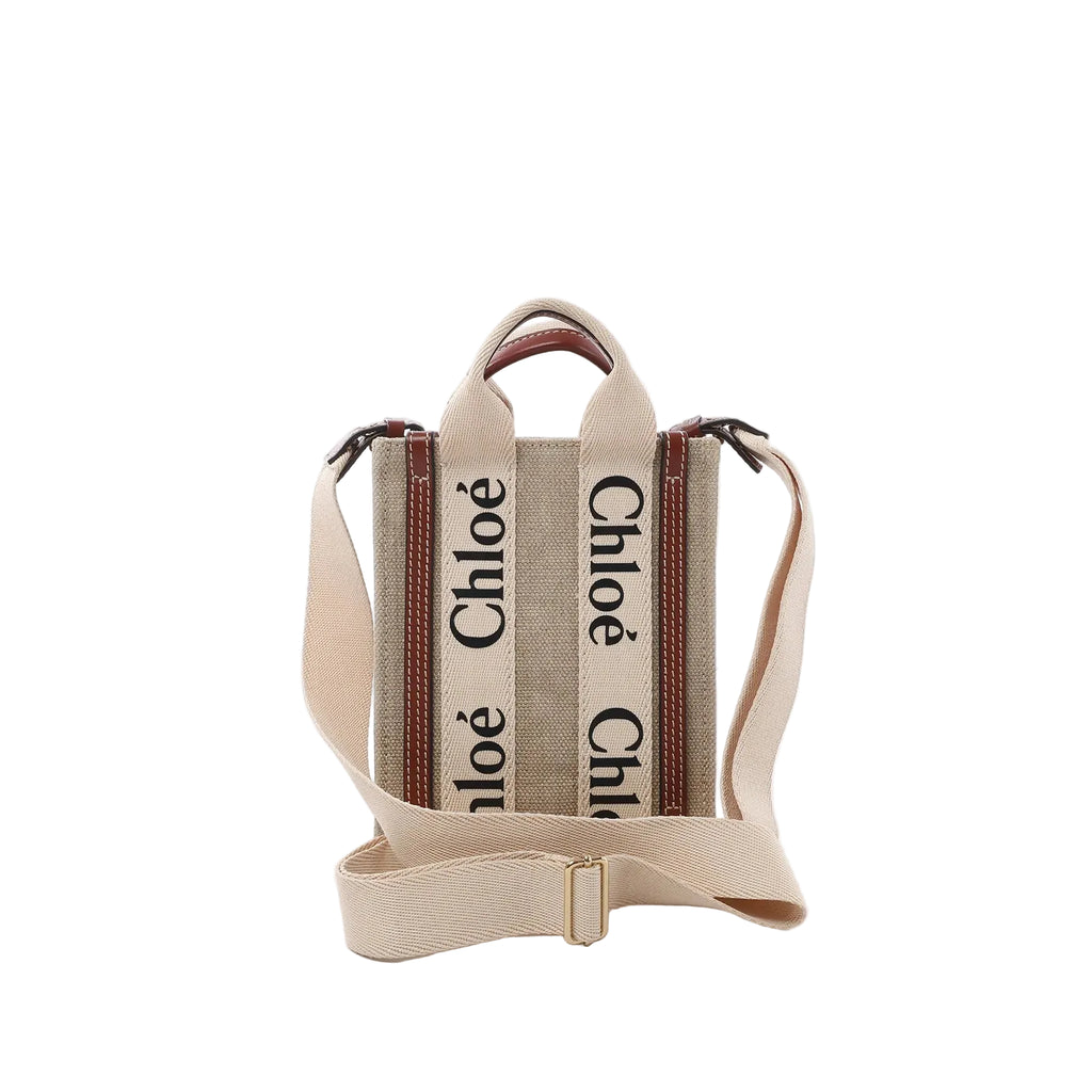 Front view of Chloé Woody North South mini tote in beige canvas with vertical “Chloé” logo webbing, brown leather trim, top handles, and an adjustable beige shoulder strap.