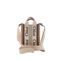 Front view of Chloé Woody North South mini tote in beige canvas with vertical “Chloé” logo webbing, brown leather trim, top handles, and an adjustable beige shoulder strap.