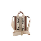 Front view of Chloé Woody North South mini tote in beige canvas with vertical “Chloé” logo webbing, brown leather trim, top handles, and an adjustable beige shoulder strap.