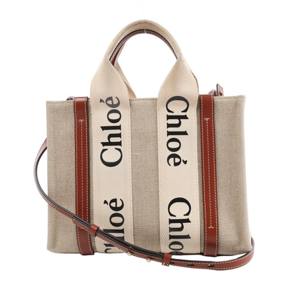 Front view of the Chloé Woody Small Canvas & Leather 2Way Handbag in beige and brown, featuring beige canvas body, brown leather trim, white stitching, logo-printed webbing top handles, and a detachable shoulder strap, displayed against a white background.