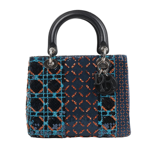 Front view of a Dior Lady Dior Medium Handbag in navy blue, embellished with orange, blue, and black sequins in geometric patterns, featuring black leather handles, silver-tone hardware, and hanging Dior letter charms, displayed against a white background.