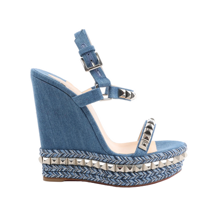 Christian Louboutin blue denim wedge sandals with silver pyramid studs on the straps, braided chevron-patterned wedge sole, and ankle buckle closure, shown in side profile on a white background.