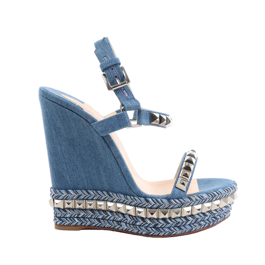 Christian Louboutin blue denim wedge sandals with silver pyramid studs on the straps, braided chevron-patterned wedge sole, and ankle buckle closure, shown in side profile on a white background.