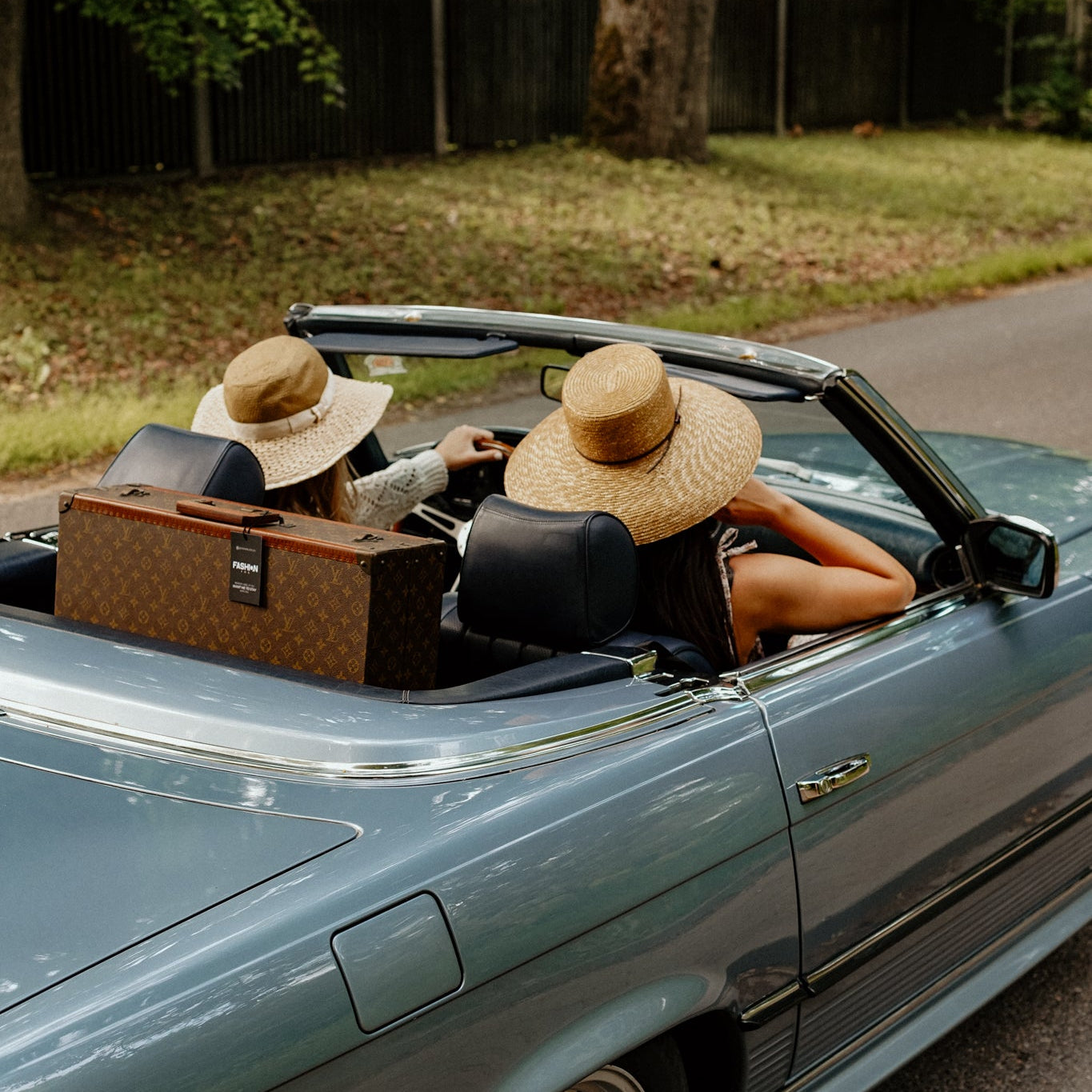 A blue convertible car parked outdoors on a road with two people wearing wide-brim straw hats sitting inside, along with a brown monogram suitcase in the back.