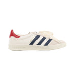 Side view of a white Gucci x Adidas Gazelle sneaker with navy stripes, a red heel tab, cream laces and an off-white sole embossed with Gucci branding.