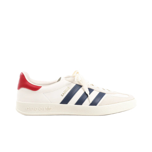 Gucci x Adidas Gazelle Leather Sneakers in white featuring smooth leather upper, navy blue three stripes, red heel tab with Adidas logo, cream laces, gold “GAZELLE” lettering, and embossed Gucci x Adidas branding on the sole, displayed on a white background.