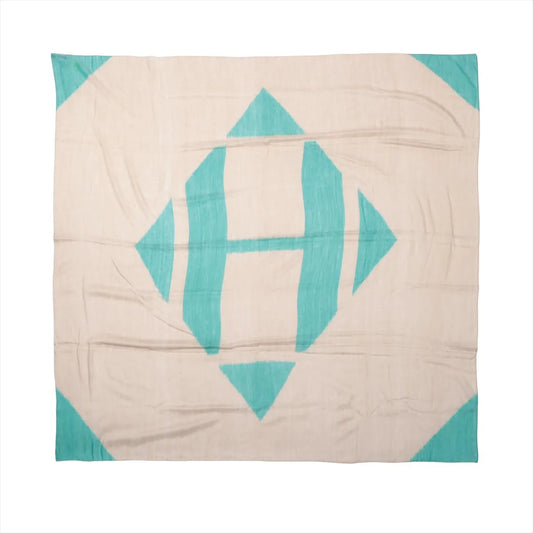 Flat-lay of a Hermès Carré Géant "Calégé Amplume HIKAT" scarf in beige featuring a large, abstract green “H” motif with geometric shapes, crafted from a lightweight cashmere and silk blend.