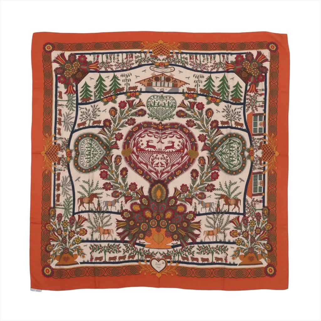 Orange Hermès Carré 140 cashmere-silk scarf laid flat with intricate multicolor folkloric motifs, floral and animal details, and a wide orange border on a white background.