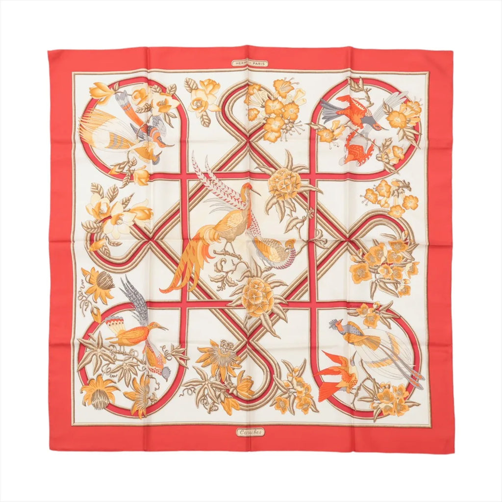 Hermès Carré 90 “Caraïbes” silk scarf with red border, ivory center, ribbon-style lines, and illustrated birds and flowers in warm tones.