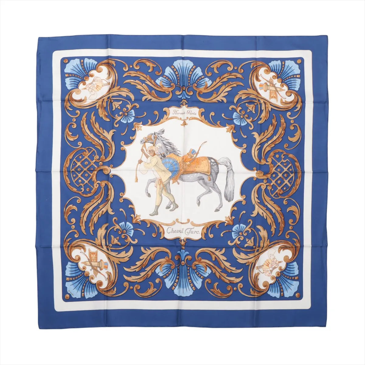 Flat-lay of a Hermès Carré 90 "Cheval Turc" silk scarf in blue featuring a central illustration of a Turkish horse and rider, surrounded by intricate baroque scrollwork and ornamental flourishes in gold, blue, and white tones.