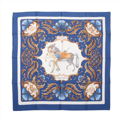 Flat-lay of a Hermès Carré 90 "Cheval Turc" silk scarf in blue featuring a central illustration of a Turkish horse and rider, surrounded by intricate baroque scrollwork and ornamental flourishes in gold, blue, and white tones.