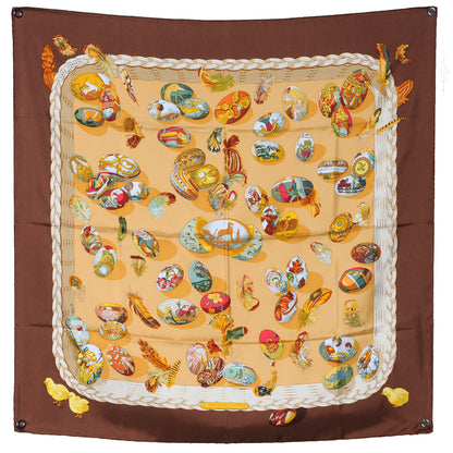 Hermès Carré 90 "Couvée d’Hermès" double-pleated silk handkerchief in brown, featuring a diamond-shaped silhouette with multicolored illustrations of birds and feathers on a warm beige background, framed by layered pleated edges.