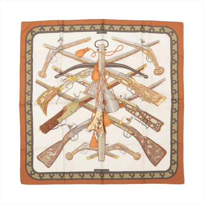 Hermès Carré 90 "Diane Gunshot" silk handkerchief in brown, featuring an ornate pattern of antique firearms and crossbows arranged symmetrically on a cream background, bordered with a burnt orange and olive green clover motif.