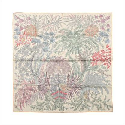 Flat-lay of a Hermès Carré Double Face 90 "Les Jardins des Peintres" silk scarf featuring a soft multicolor botanical illustration with palm leaves, flowers, and foliage in pastel tones on a cream background.