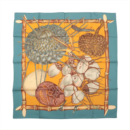 Flat-lay of a Hermès Carré 90 "Jardin Créole" silk scarf featuring a vibrant tropical composition with coconuts, woven palm, exotic fruits, and bamboo frame on a golden yellow background with a teal blue border.