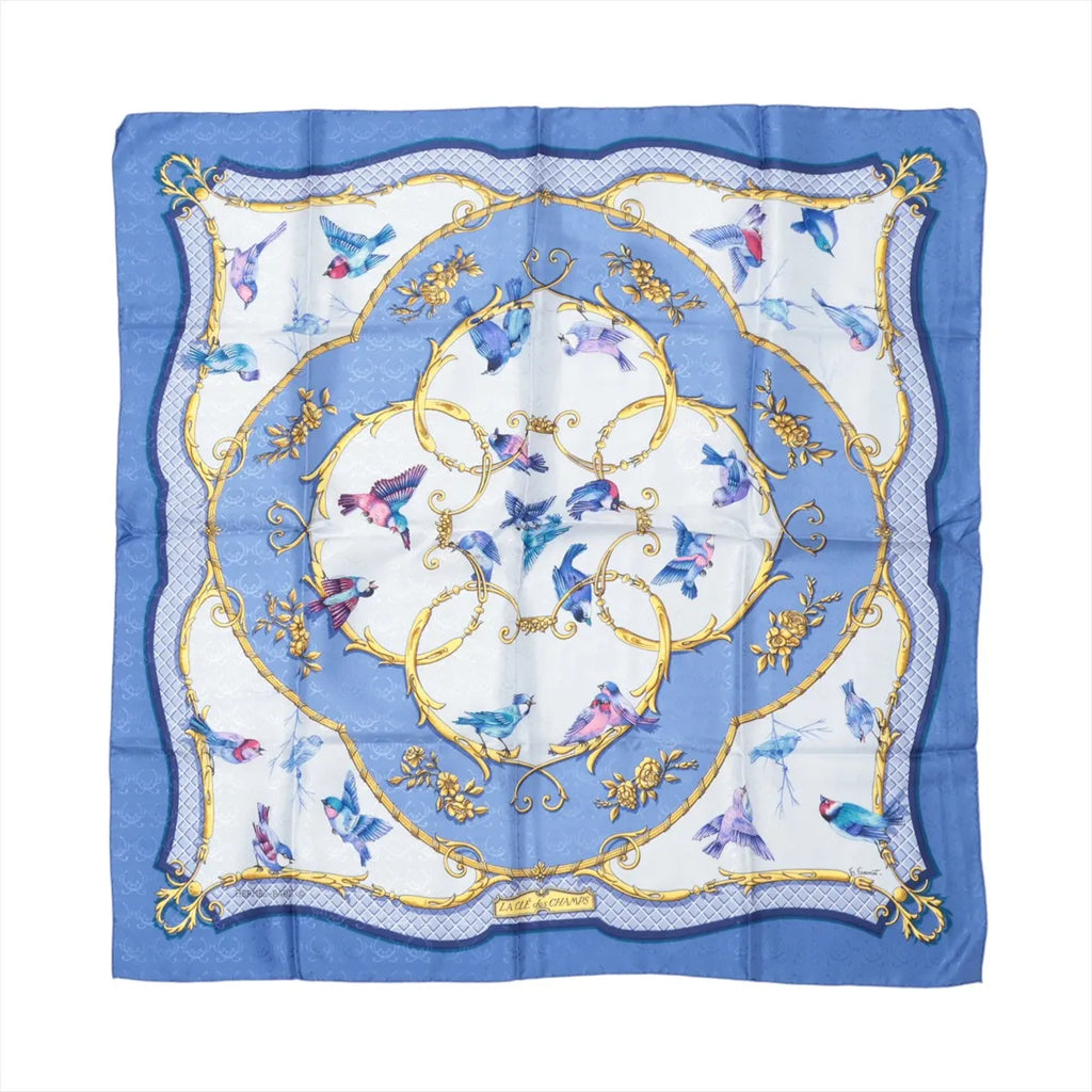 Hermès Carré 90 blue silk scarf with flying birds in blue and pink, ornate gold scrollwork circles, and a blue border with patterned corner details.