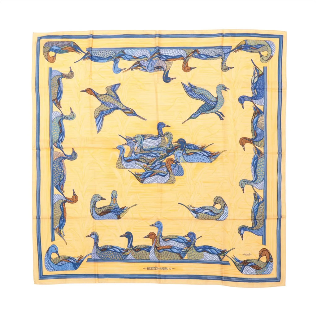 Flat-lay image of Hermès Carré 90 "La Mare aux Canards" silk scarf featuring a yellow background with an intricate duck pond motif in blue, orange, and brown tones.