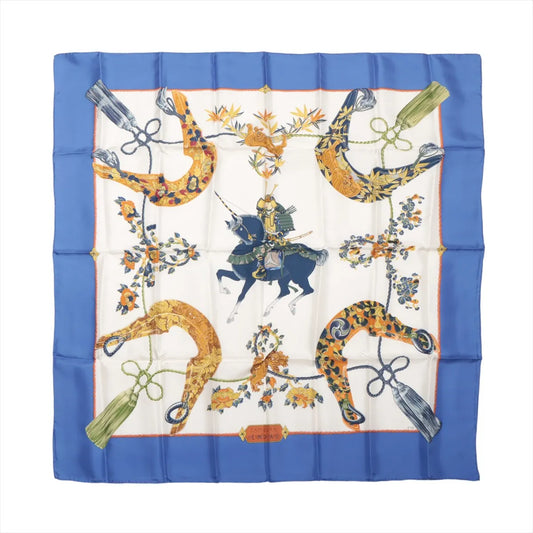 Hermès Carré 90 "SAMOURAI" silk scarf in blue with intricate samurai warrior and horse illustration at center, surrounded by golden stirrups, floral motifs, and tassels on an ivory background with a vivid blue border.